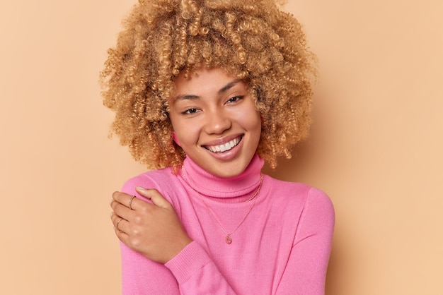 Portret van een goed uitziende vrouw glimlacht aangenaam houdt de hand op de schouder kijkt naar de camera met een vrolijke uitdrukking gekleed in roze coltrui geïsoleerd over beige achtergrond. Gezichtsuitdrukkingen concept