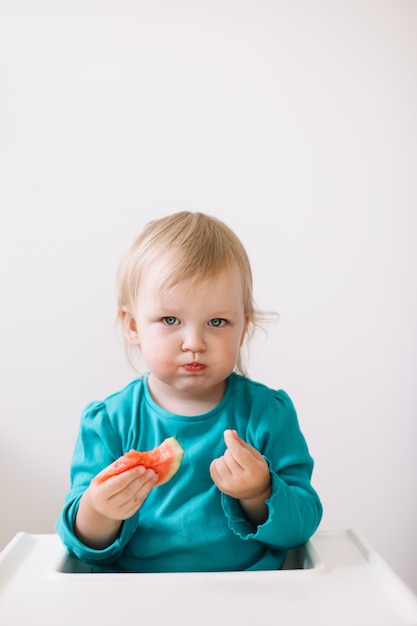 Portret van een grappig klein babymeisje dat in een kinderstoel zit en watermeloen eet