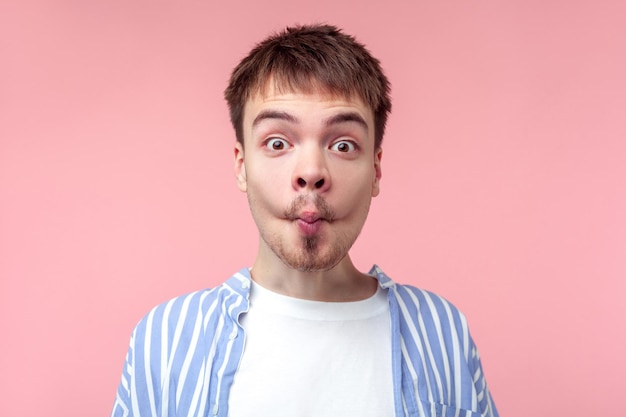 Portret van een grappige bruinharige man met een kleine baard en snor in een casual gestreept shirt die een visgezicht met lippen maakt, kinderachtig en dwaas kijkt. indoor studio-opname geïsoleerd op roze achtergrond