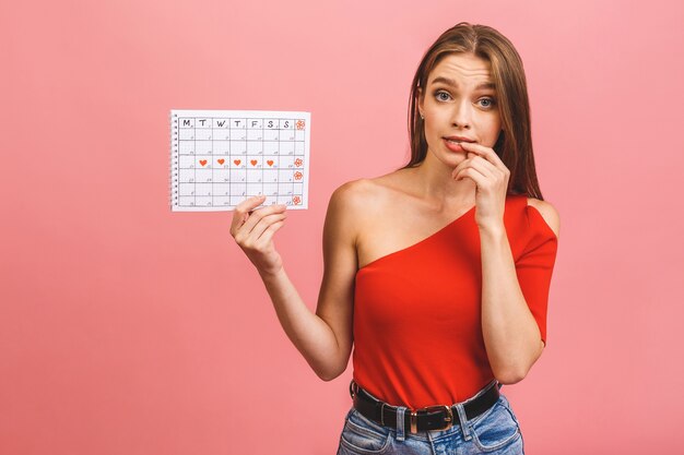 Portret van een grappige jonge de periodekalender van de meisjesholding die over roze achtergrond wordt geïsoleerd