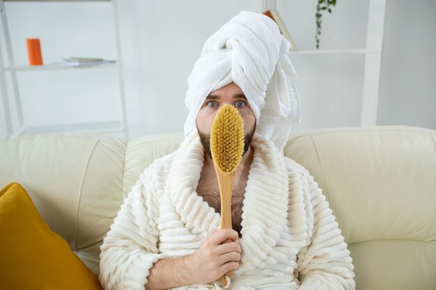 Portret van een grappige kerel die een tulbandhanddoek draagt en een massageborstel in zijn handen houdt terwijl hij op de bank zit. mannelijke huidverzorging en spa concept.