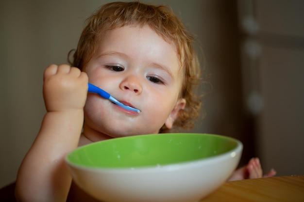 Portret van een grappige kleine babyjongen die eet van een bord dat een lepel vasthoudt, een baby die een lepel vasthoudt terwijl