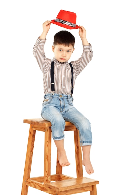 Portret van een grappige kleine jongen, zittend op een hoge kruk in een rode hoed op wit wordt geïsoleerd