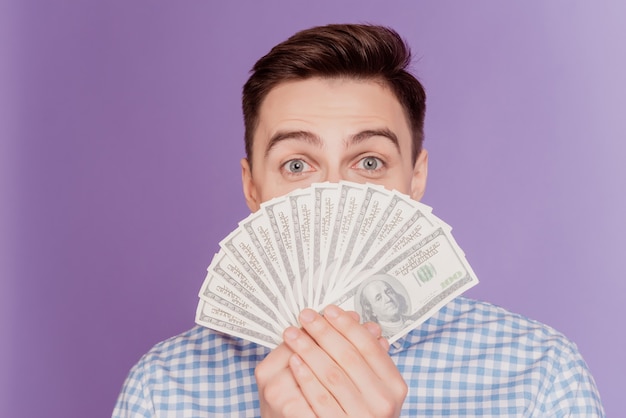 Portret van een grappige, rijke, verlegen man die contant geld houdt, een fan verbergt het gezicht op een violette achtergrond