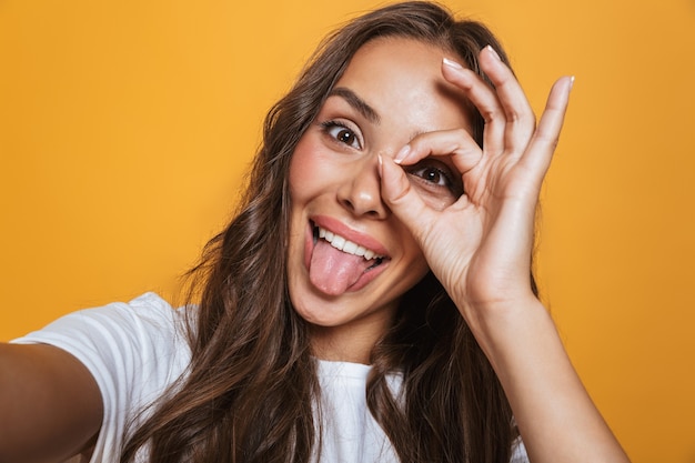 Foto portret van een grappige vrouw 20s met lang haar die plezier hebben tijdens het nemen van een selfiefoto, geïsoleerd over gele muur