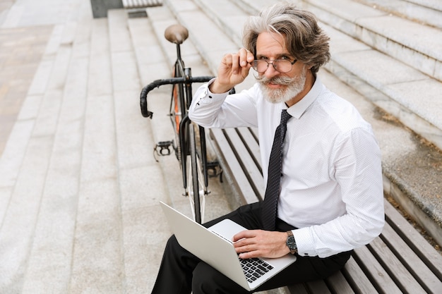 Portret van een grijsharige glimlachende zakenman in bril die op laptop typt terwijl hij op een bank zit met een fiets buitenshuis