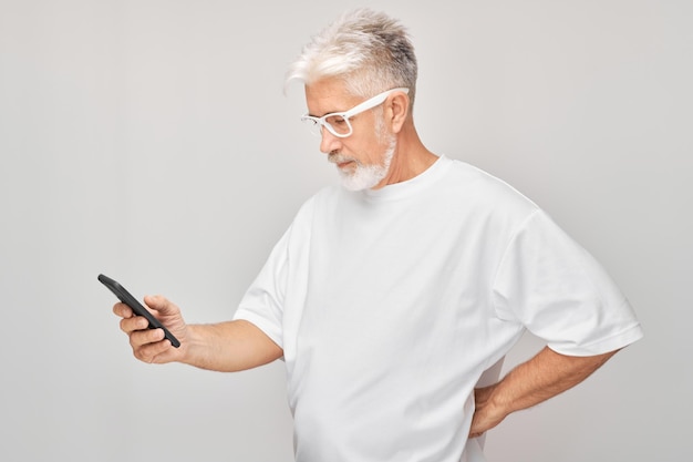 Portret van een grijsharige man in een wit T-shirt die op zijn mobiele telefoon kijkt en denkt: Persoon met een smartphone geïsoleerd op een witte achtergrond