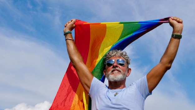 Portret van een grijsharige oudere blanke man met een baard en zonnebril die een regenboog LGBTQIA-vlag vasthoudt tegen een hemelachtergrond Biseksuele homo viert Pride Month Coming Out Day