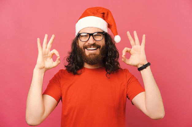 Portret van een handsone jongeman met baard die een goed gebaar toont over roze bacground en een kerstmanhoed draagt