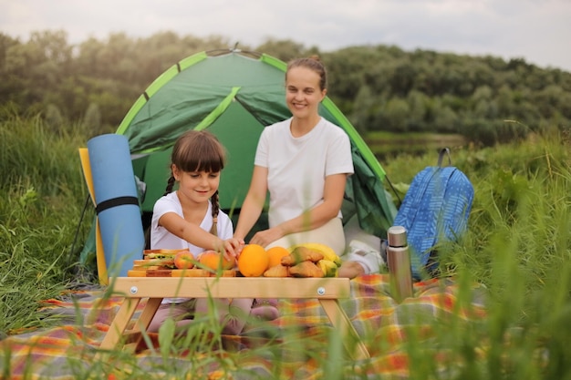 Portret van een hongerig meisje en haar moeder die op een deken bij de tent zitten en een picknickdochter hebben die eten neemt om mensen te eten die samen tijd doorbrengen en genieten