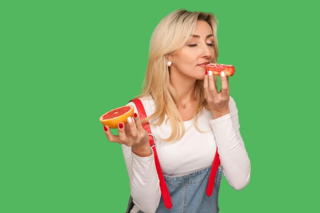 Portret van een hongerige vrouw die geniet van de geur van heerlijke zoete donut terwijl ze grapefruit in de andere hand houdt, concept van het kiezen van ongezond junkfood in plaats van fruit. studio-opname geïsoleerd op groene achtergrond