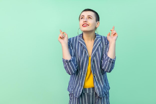 Portret van een hoopvolle knappe, mooie, stijlvolle jongedame met kort haar in een casual gestreept pak, staande, gekruiste vingers en wens om te winnen. indoor studio opname geïsoleerd op lichtgroene achtergrond.