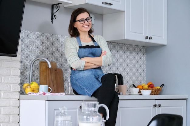 Portret van een huisvrouw in een schort thuis in de keuken, glimlachend gelukkig vrouwtje aan tafel