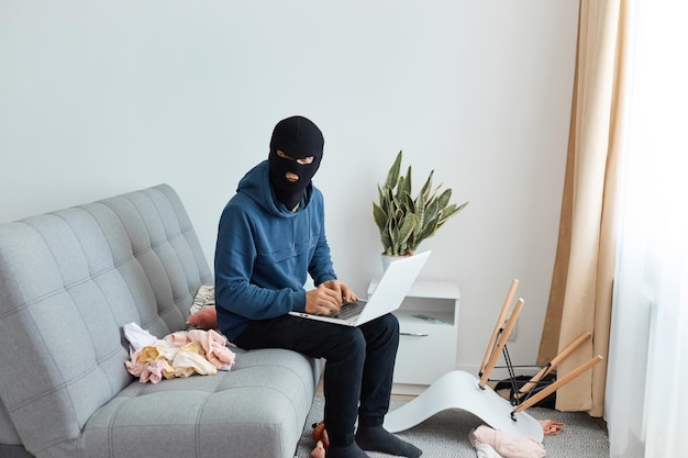 Portret van een inbreker met een blauwe hoodie en een zwart masker die hoest zit met een notitieboekje in het appartement van iemand anders en wegkijkt omdat hij bang is om door de politie te worden betrapt