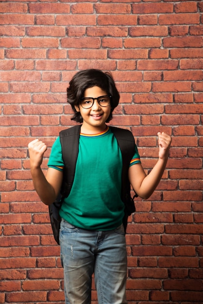 Portret van een Indiase Aziatische kleine jongen student in vrijetijdskleding met schooltas of tas, staande tegen rode bakstenen muur