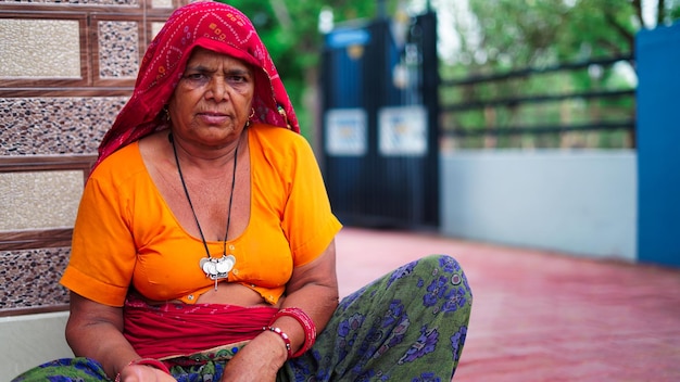 Portret van een Indiase senior vrouw die bij de hoofdingang zit en naar de camera kijkt