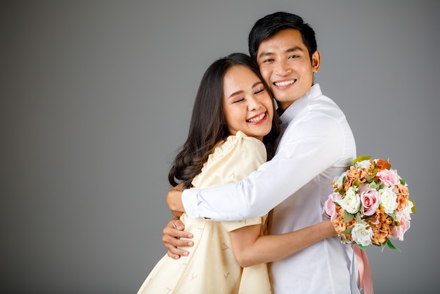 Portret van een jong, aantrekkelijk Aziatisch stel, een man met een wit overhemd, een vrouw met een beige jurk die elkaar omhelst, een vrouw met een boeket bloemen. Concept voor pre-huwelijksfotografie.