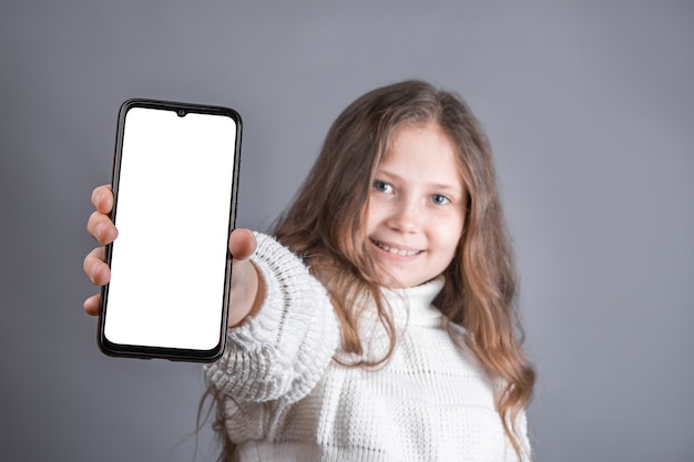 Portret van een jong aantrekkelijk meisje met blond lang golvend haar in een witte sweatergreep toont een mobiele telefoon met een wit leeg scherm op een grijze studioachtergrond. Plaats voor tekst. Kopieer ruimte.