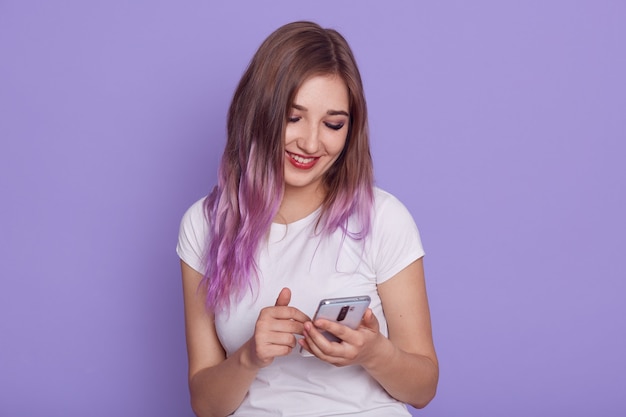 Portret van een jong aantrekkelijk meisje met violet haar dat een smartphone in handen houdt, naar het scherm kijkt met een charmante glimlach, een bericht leest, geïsoleerd op een lila achtergrond.