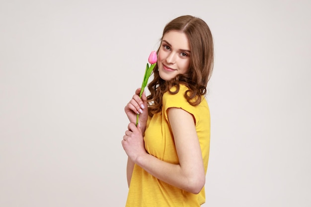 Portret van een jong bruinharig mooi tienermeisje in een geel T-shirt met tulp en glimlachend in de camera, met een romantisch cadeau. Indoor studio opname geïsoleerd op een grijze achtergrond.