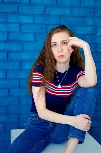 Portret van een jong en serieus meisje zittend op een stoel op een blauwe bakstenen achtergrond met lang haar in een strakke T-shirt en spijkerbroek