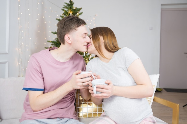 Portret van een jong gelukkig stel dat thuis ontspant met mokken in hun handen. Kersttijd en baby wachten.