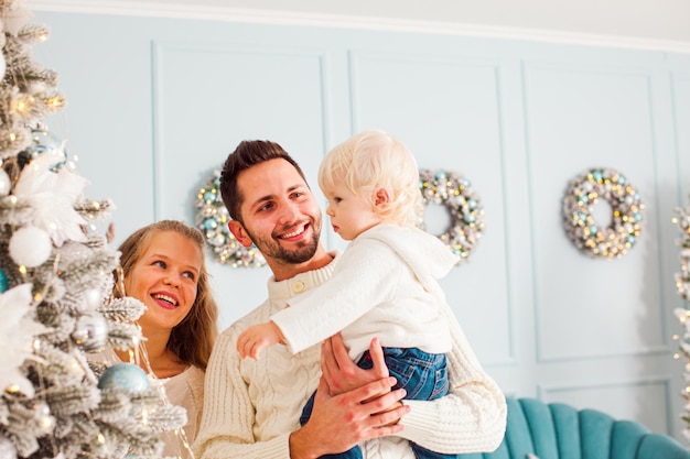 Portret van een jong gezin opknoping op decoratie op een dennenboom in hun moderne stijlvolle huis kamer of studio met trendy stijlvol interieur in blauwe en witte kleuren op een achtergrond