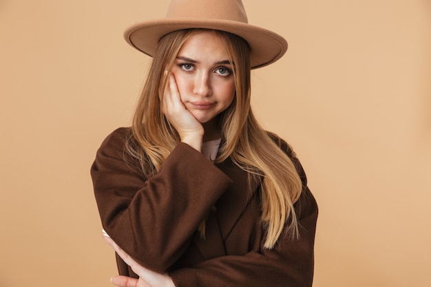 Portret van een jong kaukasisch meisje met een hoed en jas die haar hoofd omhoog steekt met een droevige blik geïsoleerd op beige