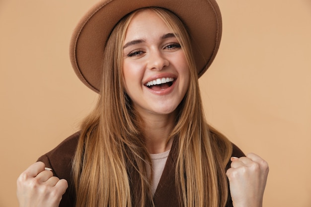 Portret van een jong kaukasisch meisje met hoed en jas glimlachend en balde haar vuisten met succes geïsoleerd op beige