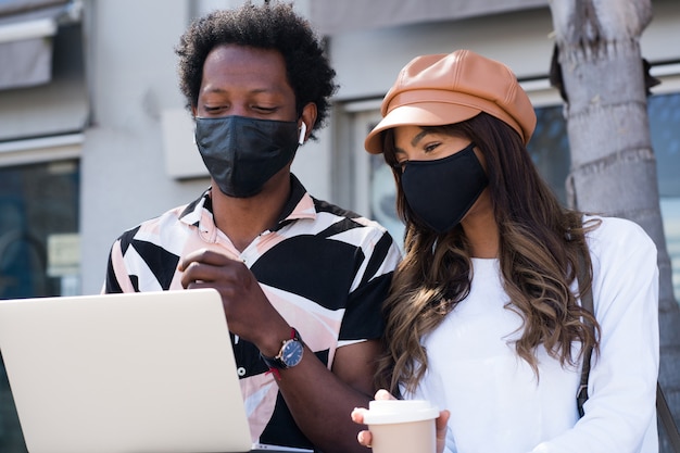 Portret van een jong koppel dat beschermend masker draagt en laptop buiten op straat gebruikt. Stedelijk concept. Nieuw normaal levensstijlconcept.