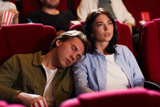 Portret van een jong koppel in de bioscoop, focus op vrouw kijken naar film met verveeld vriendje slapen op haar schouder, kopie ruimte