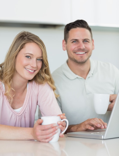 Portret van een jong koppel met koffiemokken en laptop zittend aan tafel in de keuken