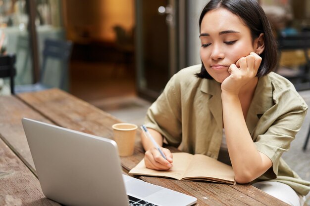 Portret van een jong koreaans meisje dat aantekeningen maakt terwijl ze online vergaderlezing luistert terwijl ze naar het laptopscherm kijkt