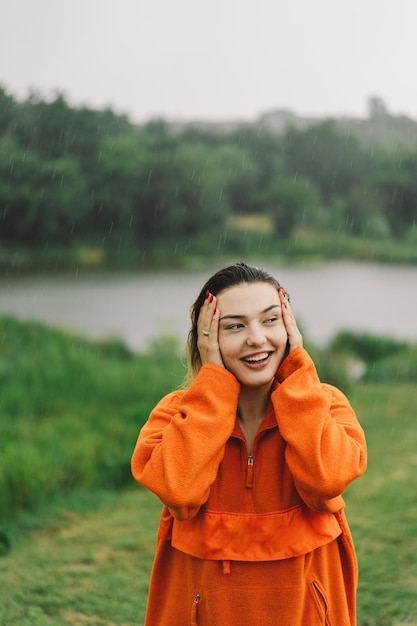 Portret van een jong meisje in een oranje trui die geniet van de regen op een zomerdag Het meisje heeft plezier en danst onder de zomerregen