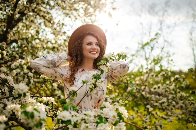 Portret van een jong meisje met donker haar dat lacht en plezier maakt in het midden van een bloeiende boom omringd door veel witte bloemen. vrouw in beige hoed en witte jurk in het lentepark.