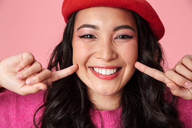 Portret van een jong mooi Aziatisch meisje met een baret die haar glimlach toont met vingers geïsoleerd op roze