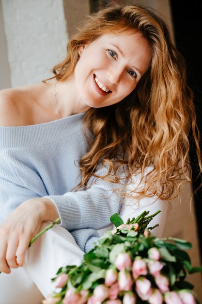 Portret van een jong mooi meisje met een boeket rozen dichtbij venster. Krullend stromend rood haar.