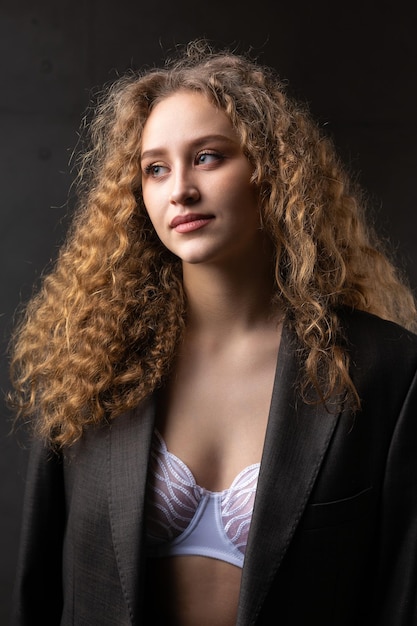 Portret van een jong roodharig krullend sexy meisje in een jas met lang haar in de studio pronkt met haar gewelfde borsten in een beha