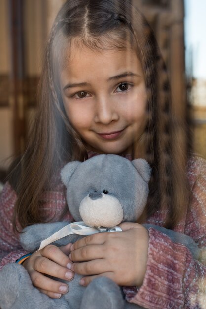 Portret van een jong schattig klein meisje dat door het raam met teddybeer kijkt