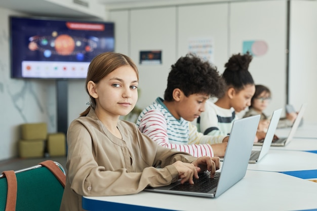 Portret van een jong schoolmeisje dat naar de camera kijkt tijdens het gebruik van een laptop in de IT-klasse, kopieer ruimte