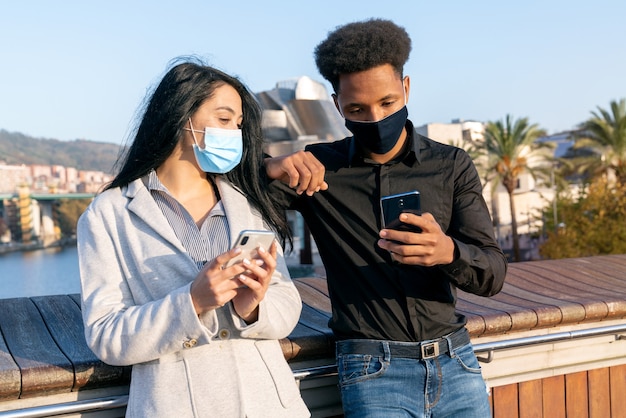 Portret van een jong stel op straat die hun mobiele telefoon gebruiken om tekst te schrijven met een gezichtsmasker vanwege de 2020 Covid-19 coronavirus pandemie jongen met afro-stijl haar