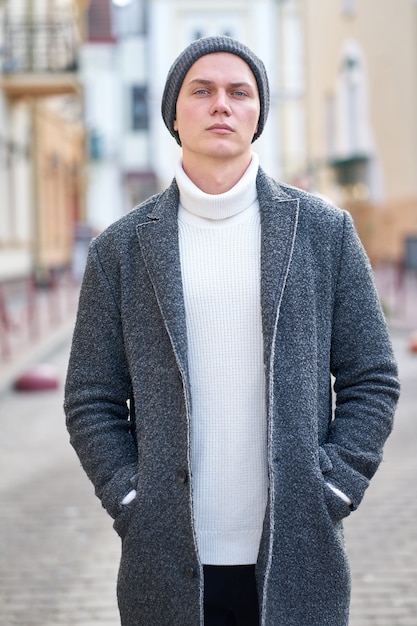 Foto portret van een jonge aantrekkelijke hipster-man met een grijze jas, een witte trui en een zwarte spijkerbroek