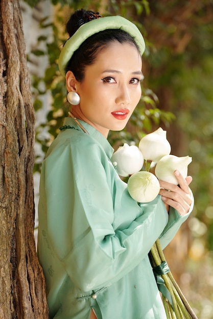 Portret van een jonge, aantrekkelijke Vietnamese vrouw in een lichtgroene traditionele kleding die witte lotusbloemen vasthoudt en naar de camera kijkt