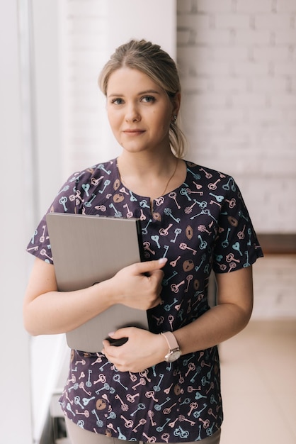 Portret van een jonge aantrekkelijke vrouw die casual kleding draagt en mappen vasthoudt terwijl ze in een lichte kantoorruimte bij het raam staat, kijkend naar de camera.