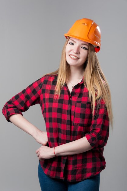 Portret van een jonge aantrekkelijke vrouw met blond haar in oranje helm en in een geruit overhemd op een neutrale grijze achtergrond.