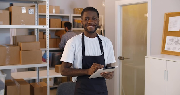 Portret van een jonge Afro-Amerikaanse man die een digitale tablet gebruikt in het magazijn en inventariseert terwijl hij naar de camera glimlacht