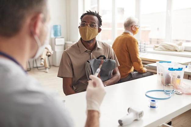 Portret van een jonge Afro-Amerikaanse man die een masker draagt tijdens het wachten op vaccinatie in de kliniek, kopieer ruimte