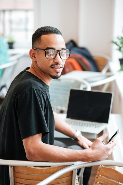 Portret van een jonge afro-amerikaanse student met een bril die een mobiele telefoon vasthoudt en naar voren kijkt terwijl hij in een café zit