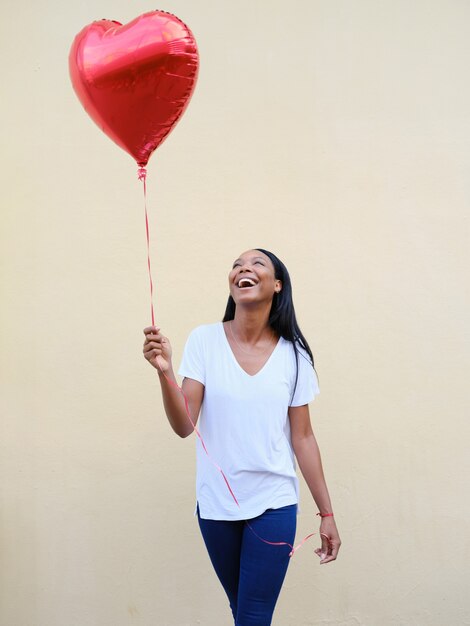 Portret van een jonge Afro-Amerikaanse vrouw die een hartvormige ballon vasthoudt terwijl ze buiten tegen een muur staat. Liefdesconcept.