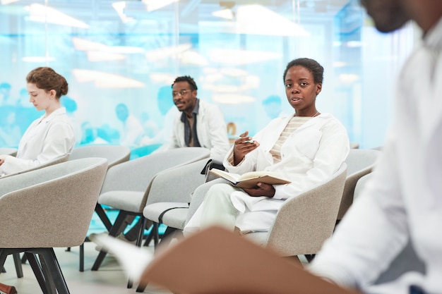 Portret van een jonge afro-amerikaanse vrouw die een laboratoriumjas draagt terwijl ze in het publiek zit en luistert naar een lezing over medicijnen op de universiteit of een coworkingcentrum, kopieer ruimte